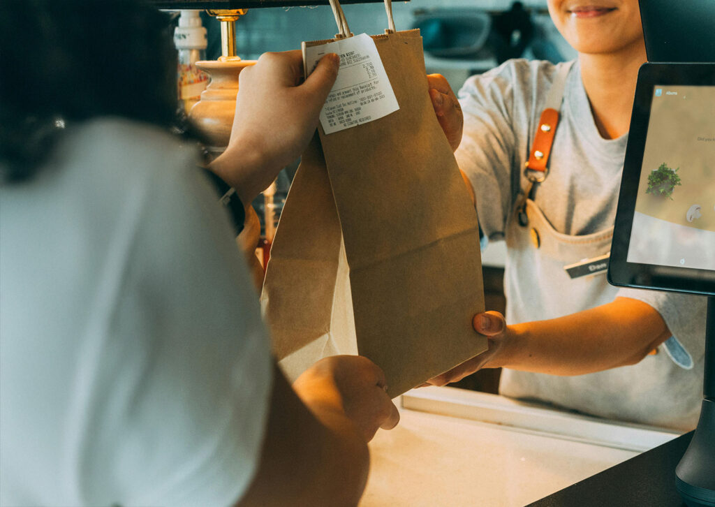 A cafe employee hands the customer's order to them in a large paper bag